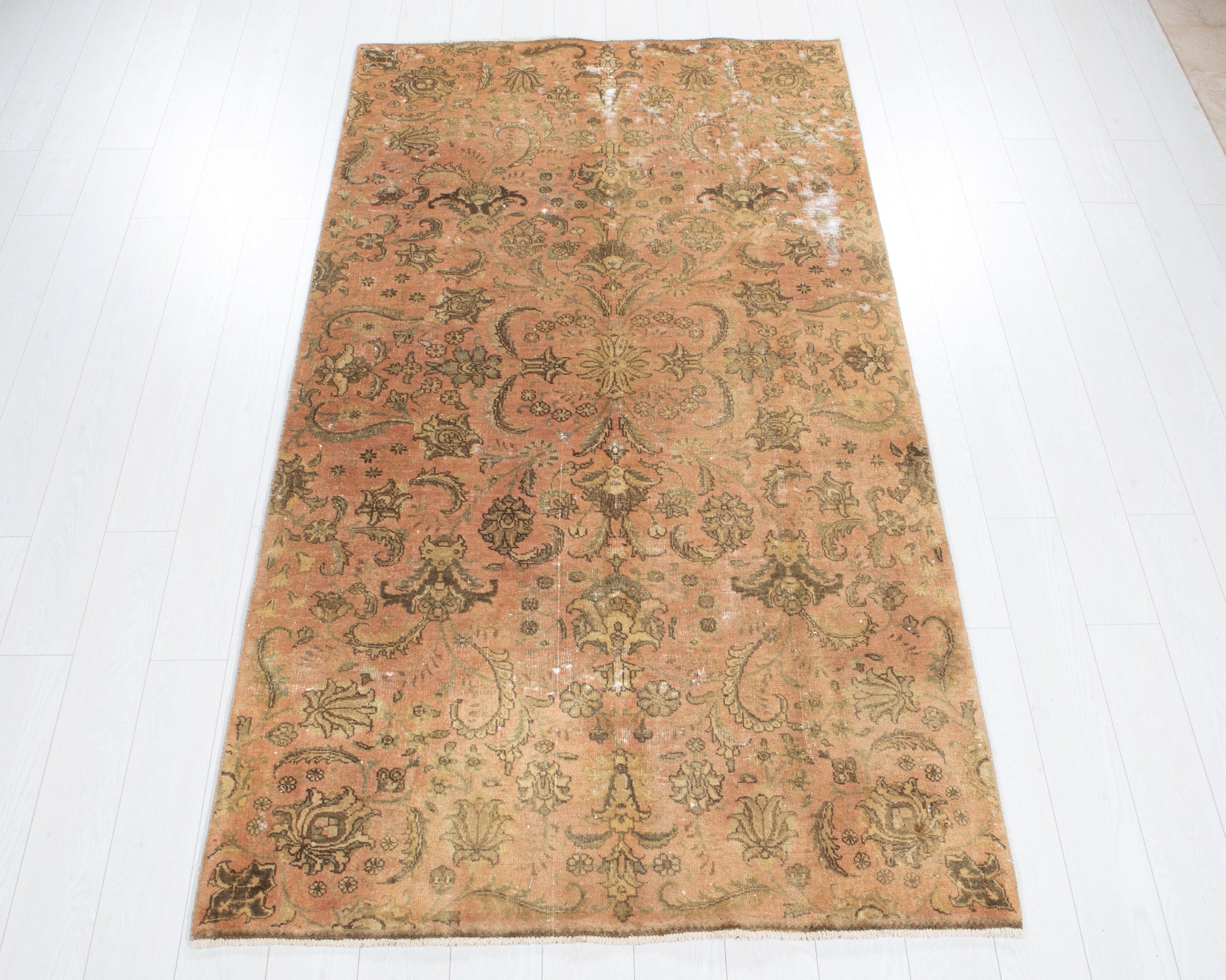 A rectangular vintage-style area rug with an ornate floral and medallion pattern in faded peach, tan, olive green, and brown tones, laid flat on a white plank floor.