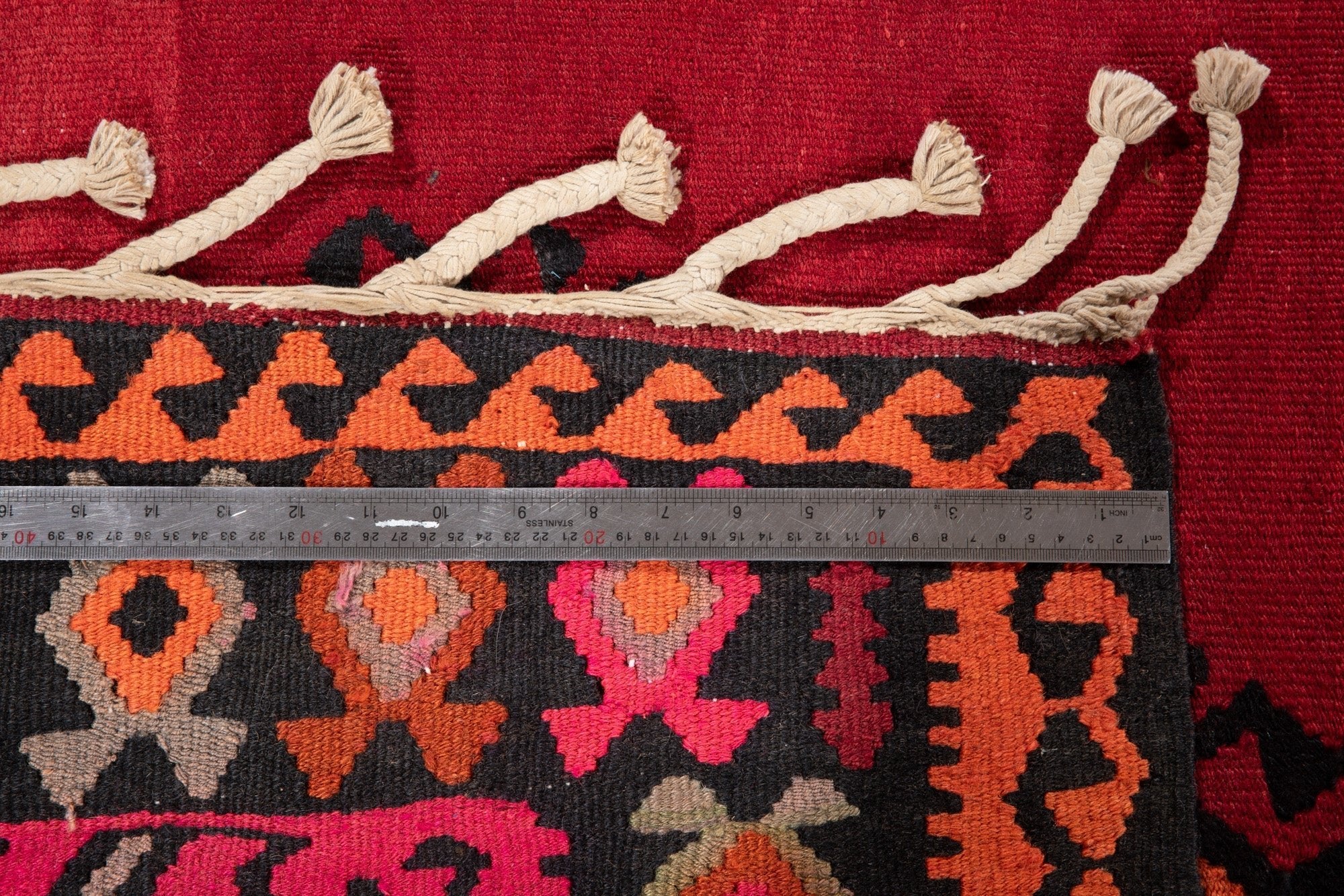 Ruler measuring the kilim's border width next to its braided fringe ties.