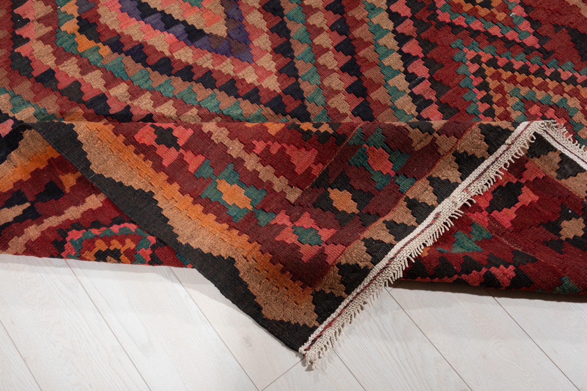 Corner of the kilim rug folded up, showing its diamond pattern and fringed edge.