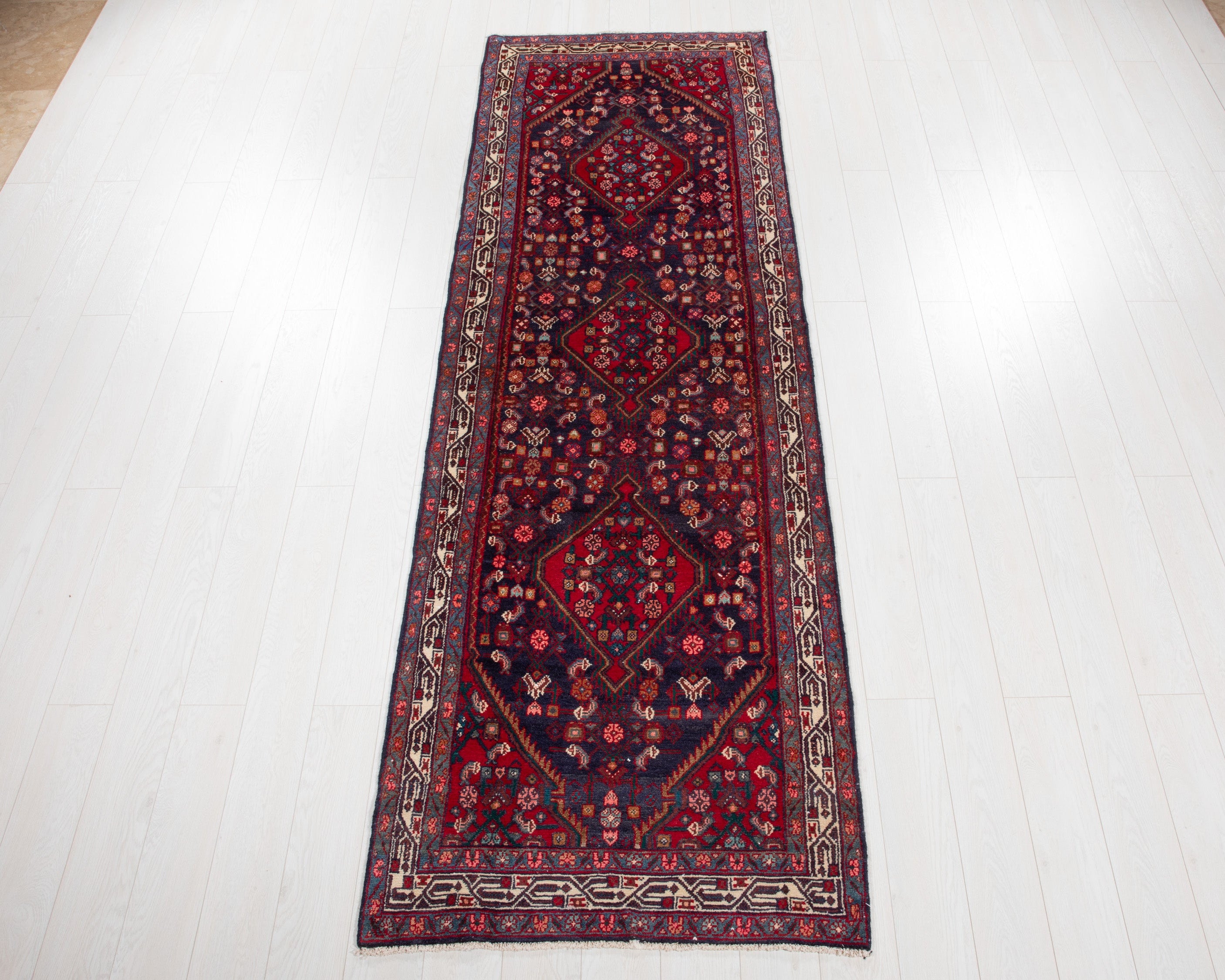 A long rectangular Persian-style runner rug with a dark red and navy blue central field, intricate floral motifs, and a patterned border, laid out on a white wooden plank floor.