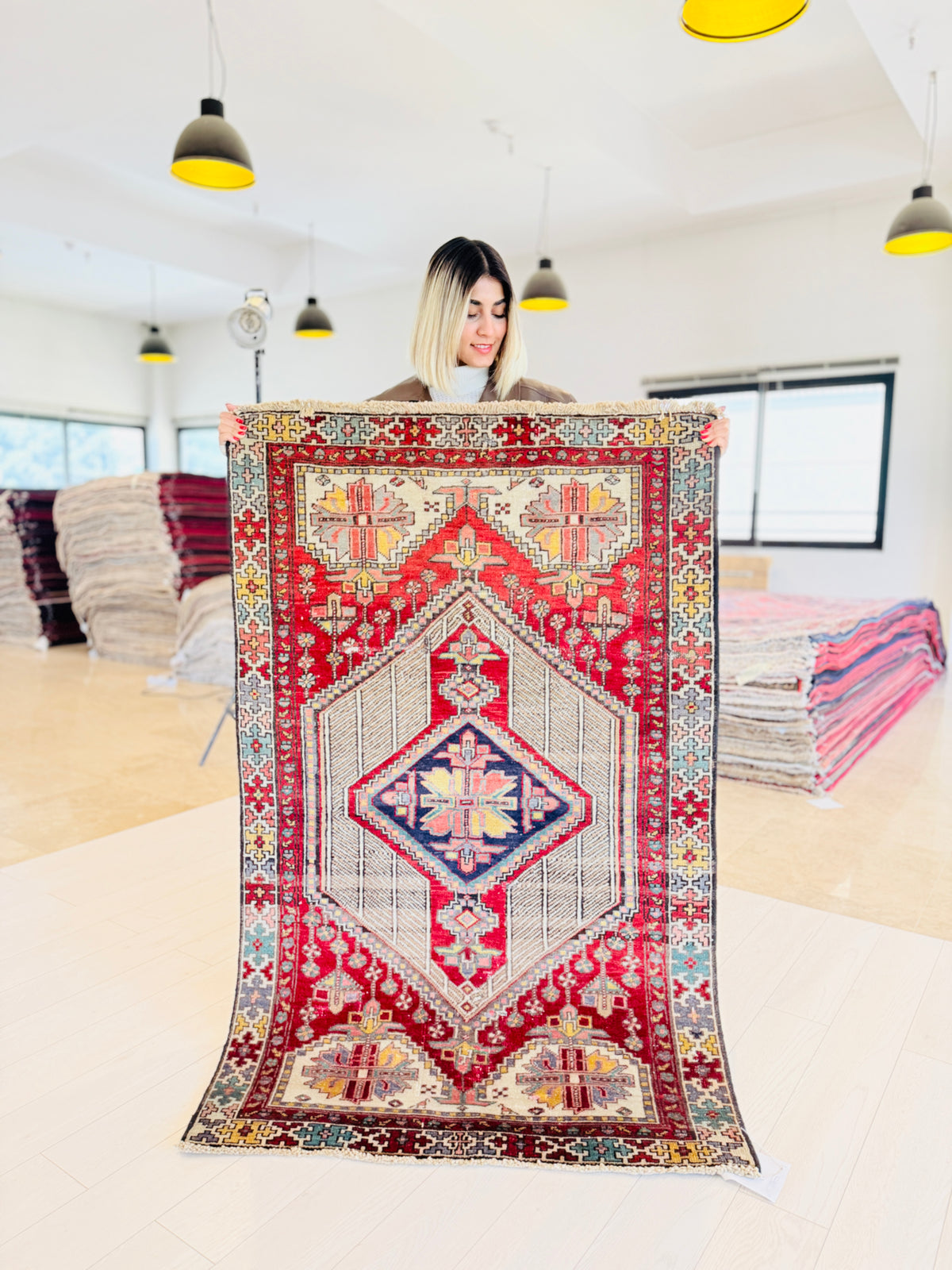 A woman holds up a large, traditional Persian-style area rug with a vibrant red, blue, and beige geometric pattern in a bright, modern room with hanging pendant lights.
