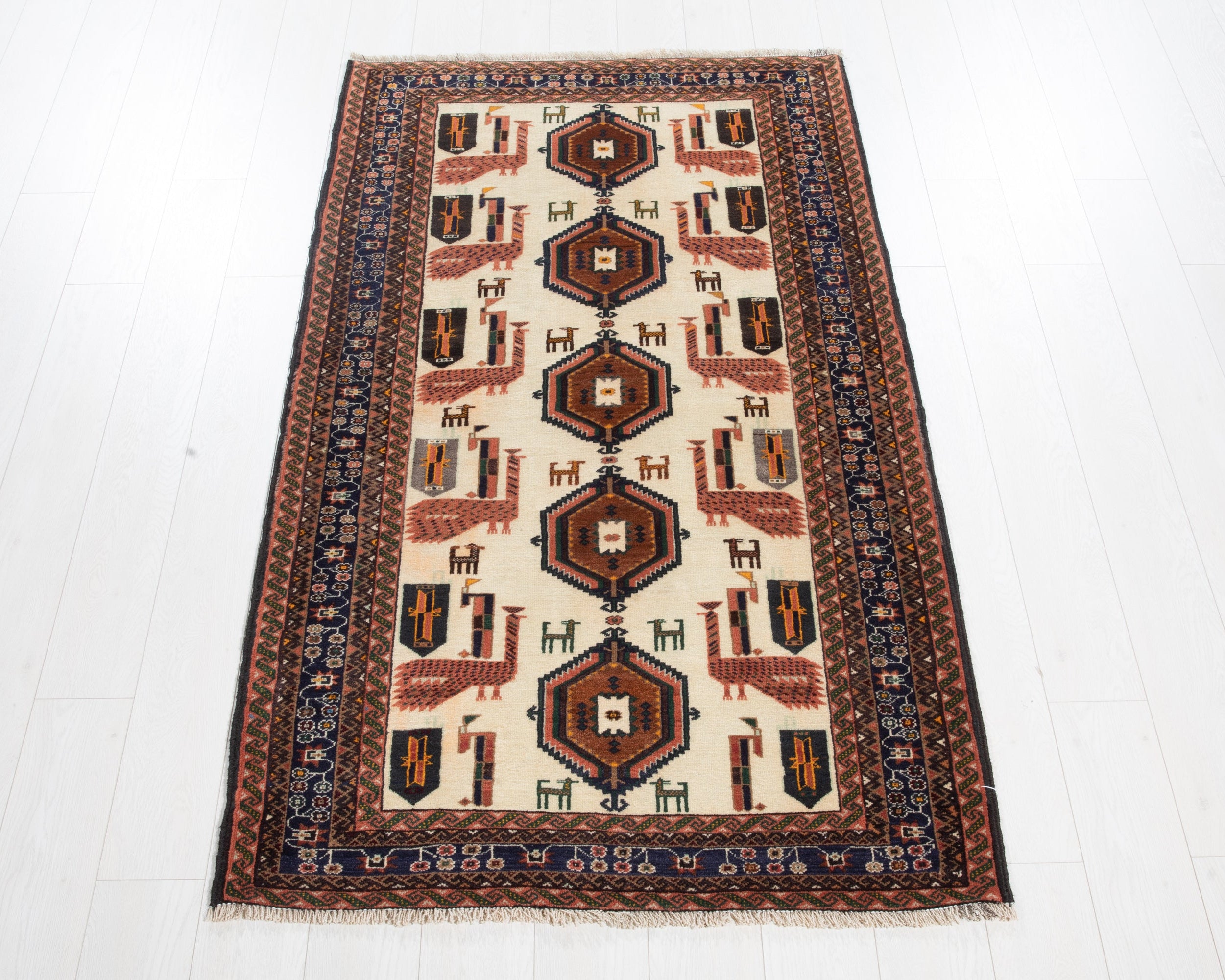 A traditional rectangular area rug with a cream center field, featuring a repeating geometric pattern of dark blue, rust red, and brown motifs, finished with a detailed dark blue and rust border.