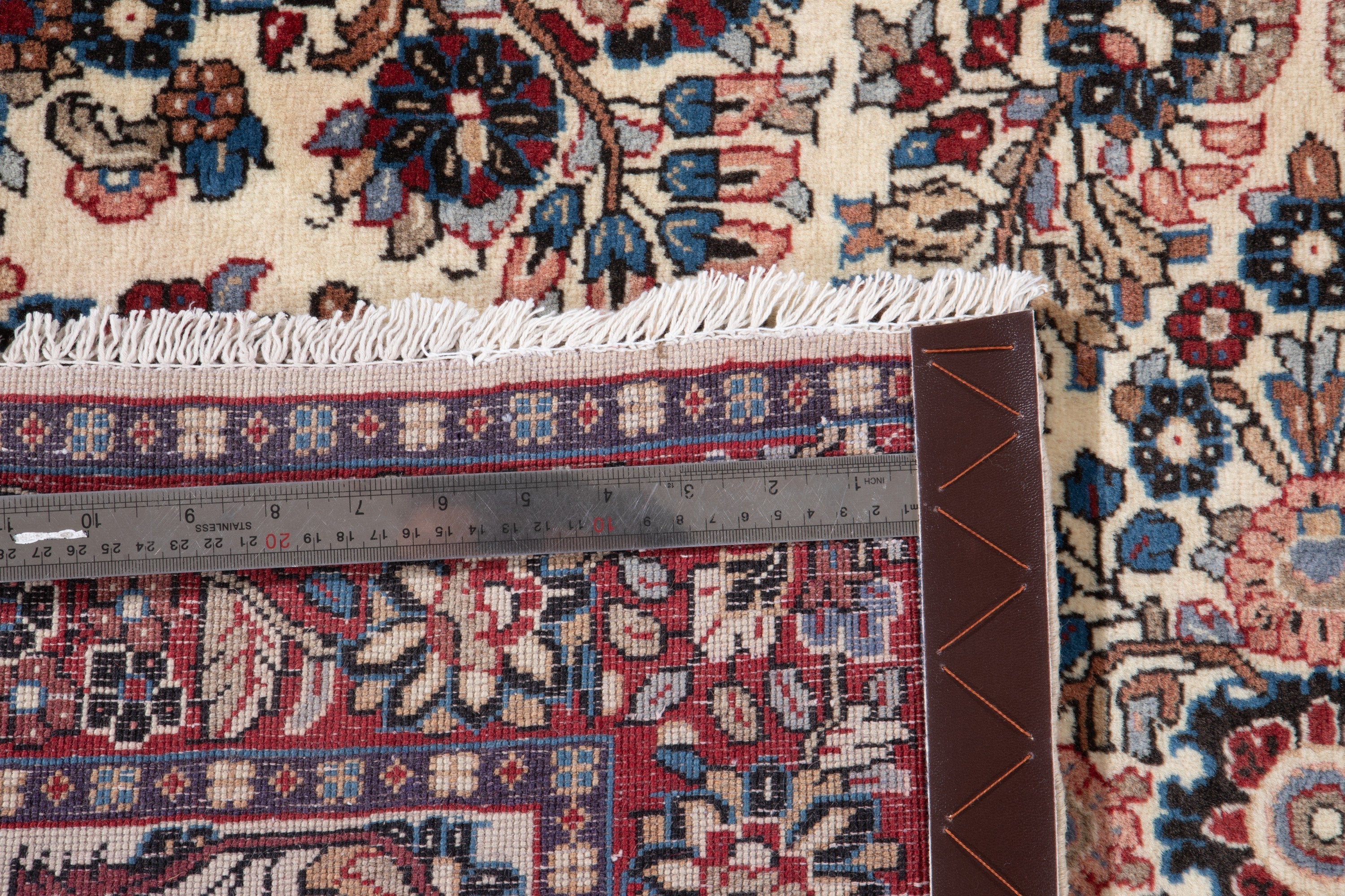 Measuring tape on the rug's edge with a leather item for scale, showing fringe and border detail.