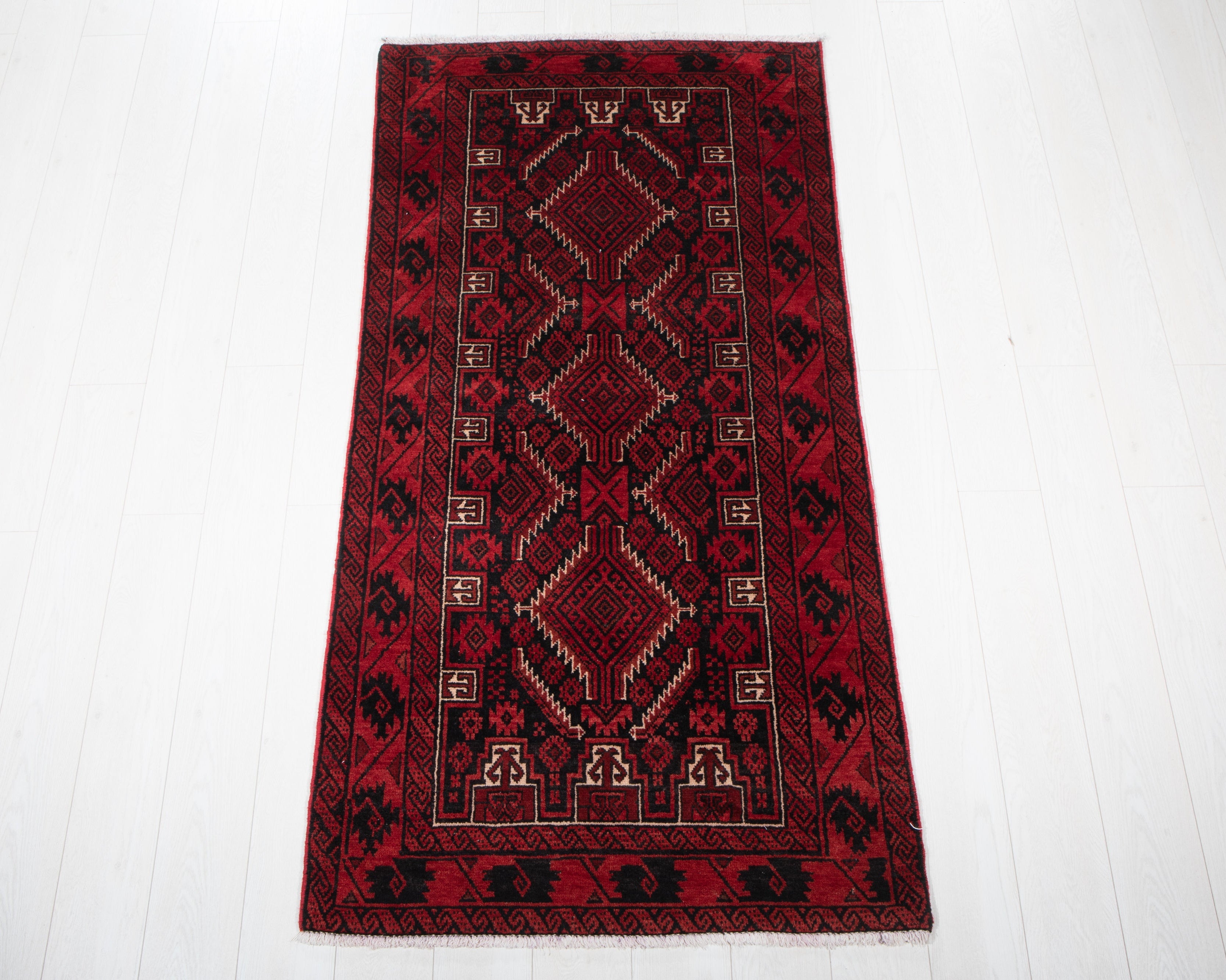 A traditional rectangular area rug with a deep red field, featuring a central diamond pattern in black, beige, and red, and a matching red border, laid out on a white wooden floor.