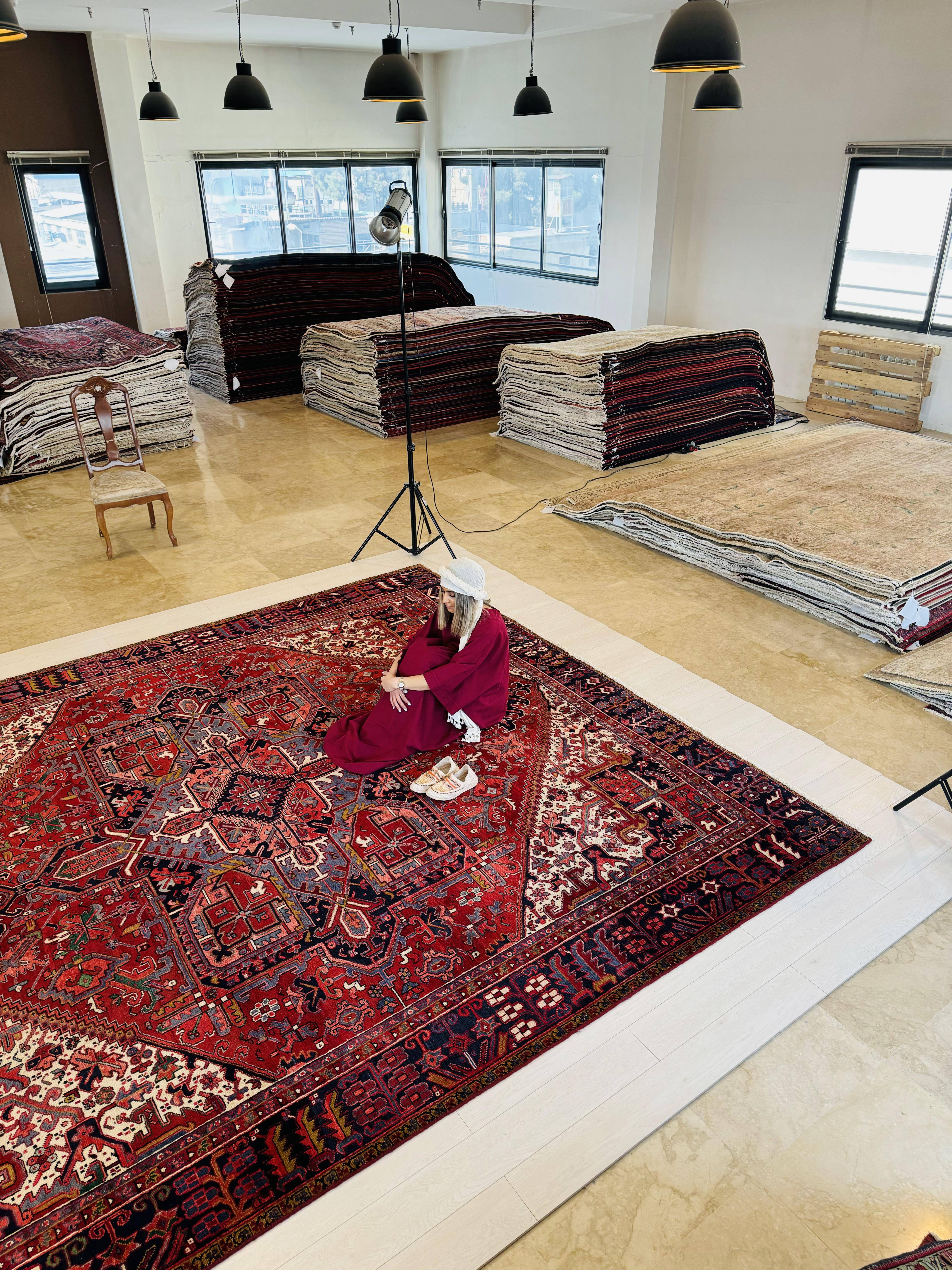 Wide showroom shot of the rug with a person sitting on it beside stacks of other rugs.