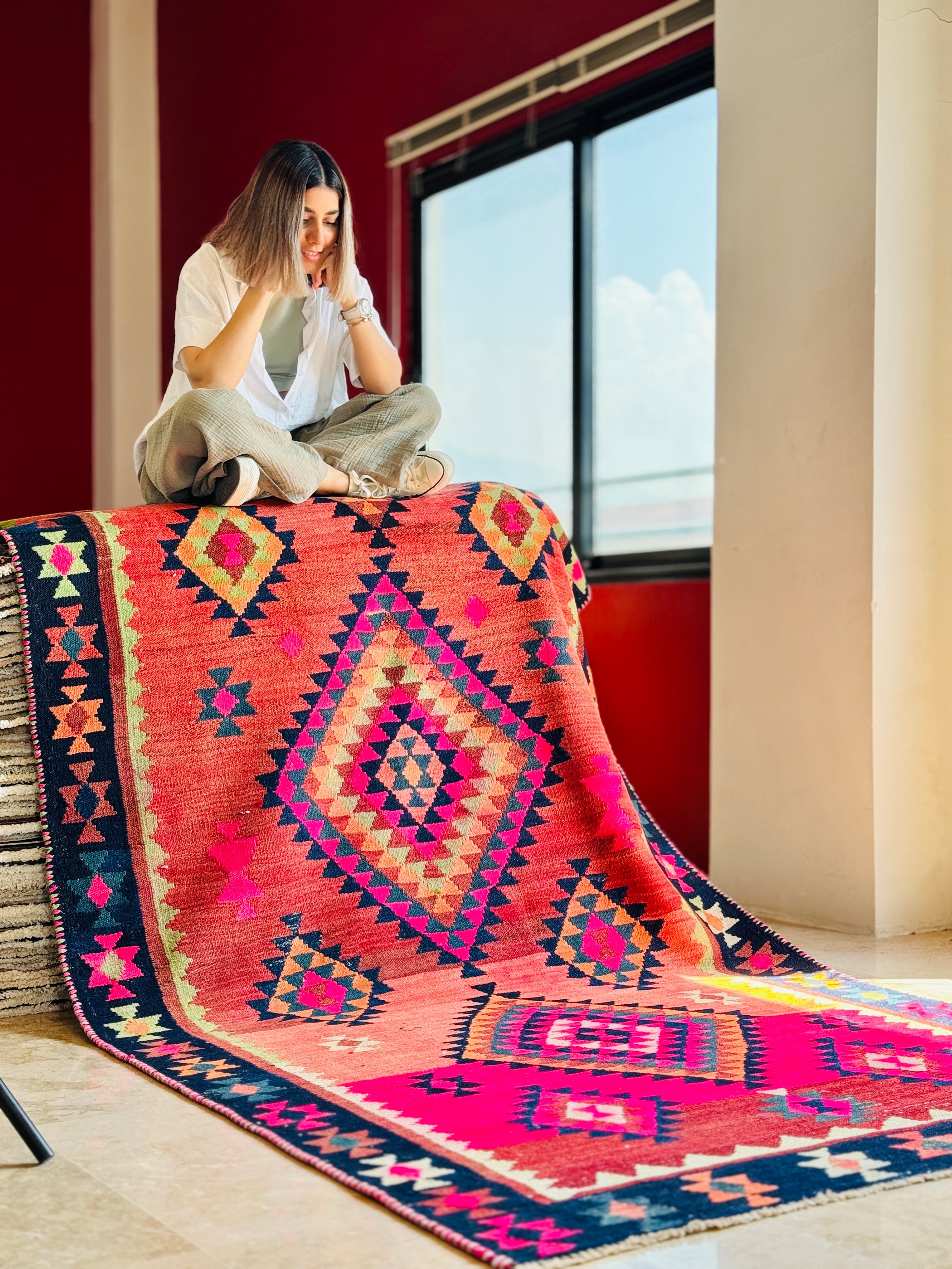 Lifestyle shot of a woman sitting on the red kilim rug draped over furniture by a window.
