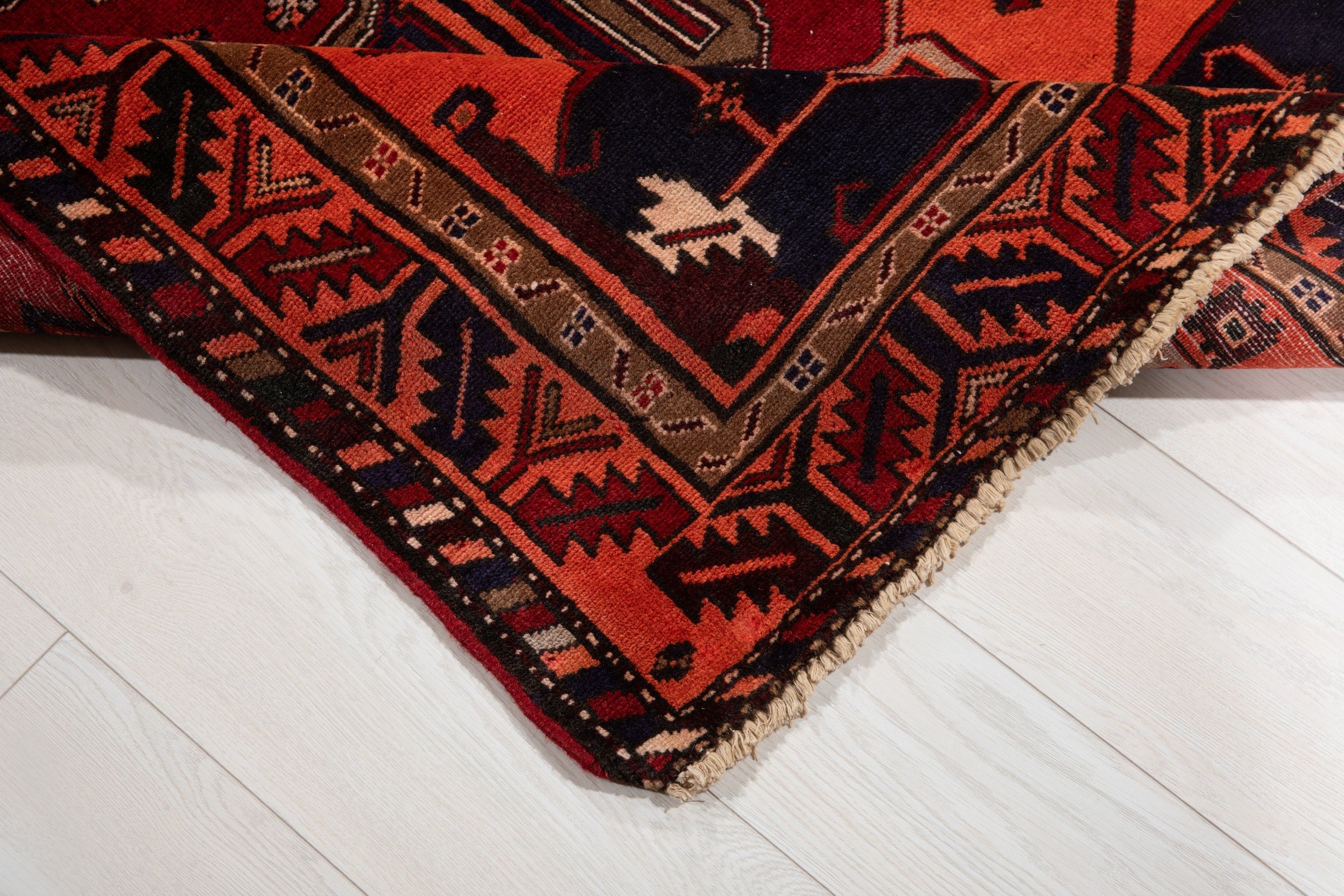Corner of the rug folded up on a wood floor, showing the border pattern and fringe.