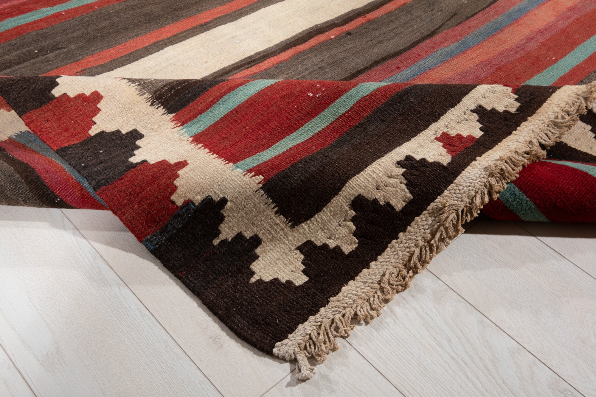 Corner of the kilim rug folded up, showing its zigzag border and fringed edge.