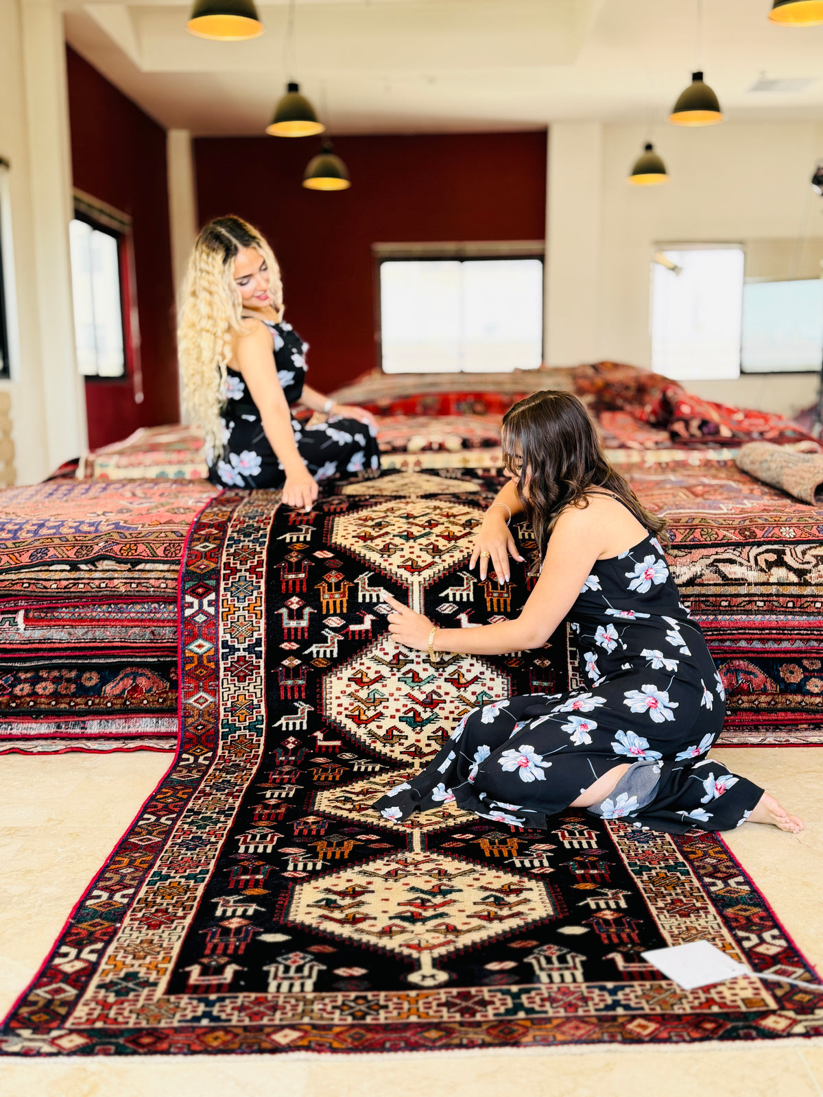 Two women in matching black floral dresses examine a large, detailed oriental area rug spread across a room with other rugs in the background.