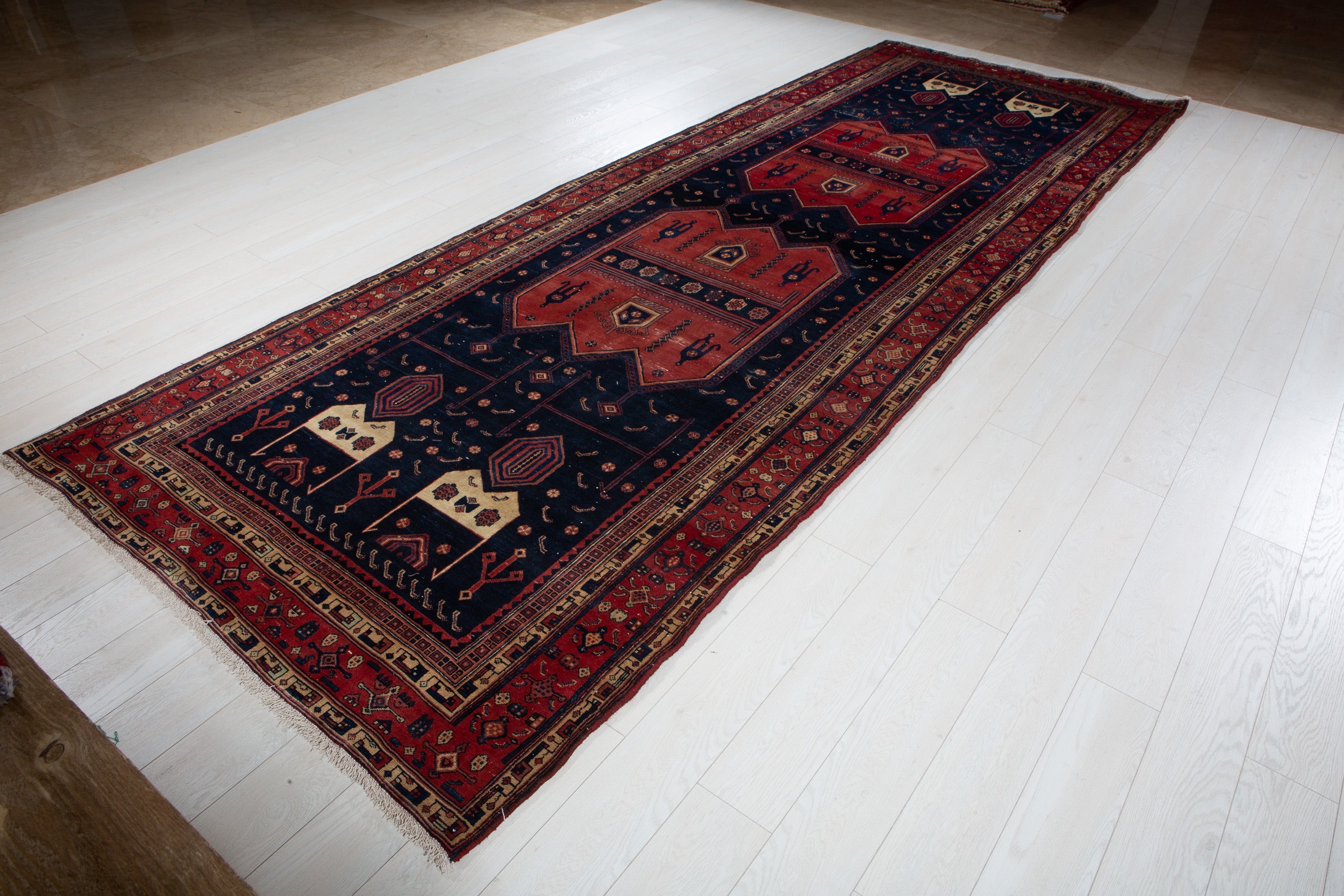 Angled overhead view of the full Kurdish Persian rug on a wood floor, showing the complete red and navy pattern.