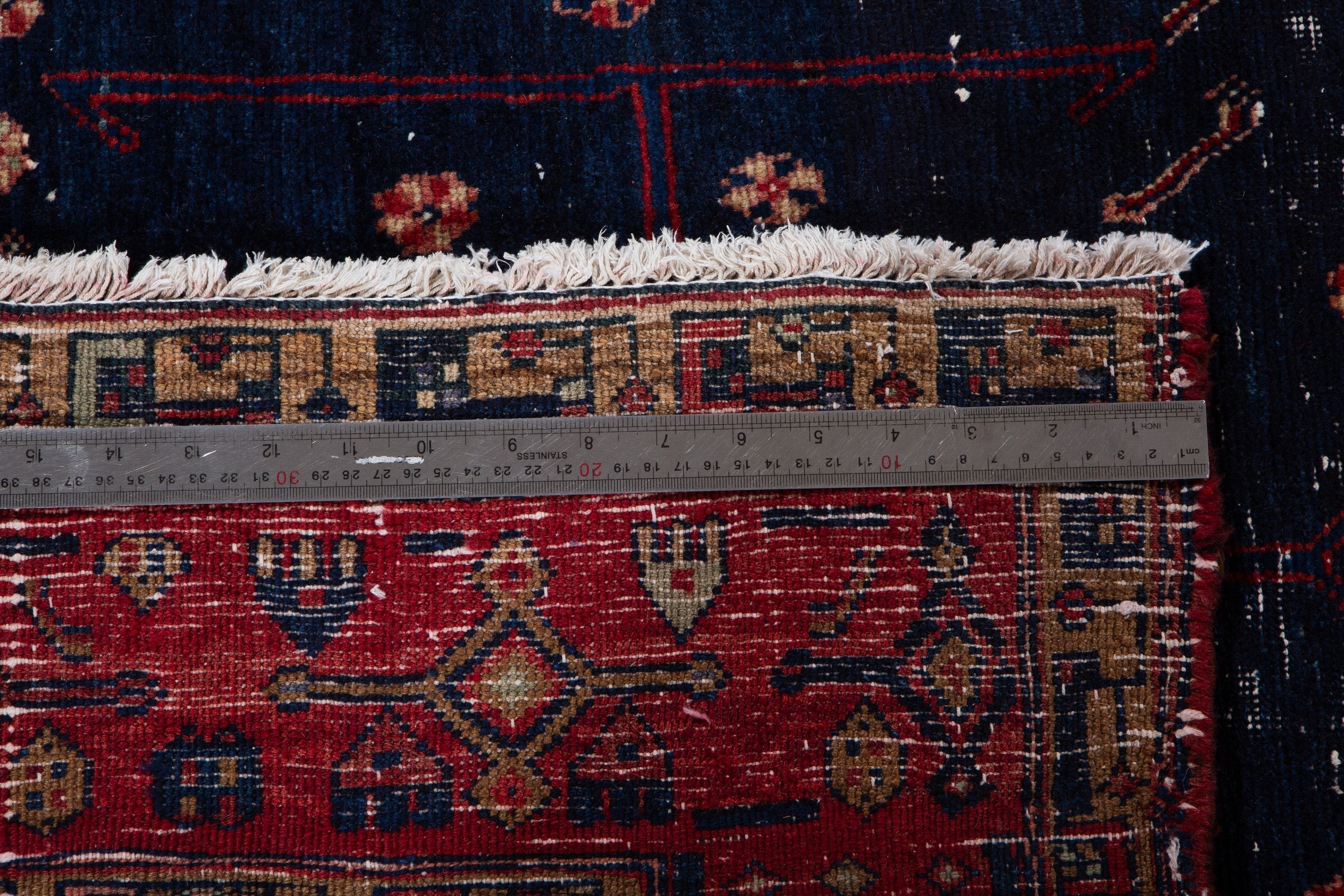 Close-up with a measuring tape across the rug's edge, showing fringe and border pattern detail.