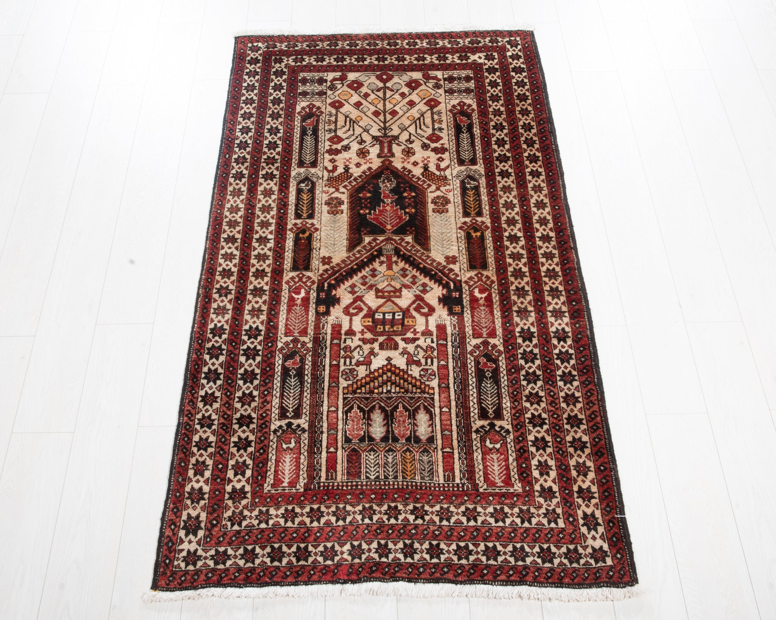 A traditional rectangular Islamic prayer rug with an intricate geometric and floral pattern in red, beige, and black, laid flat on a white wooden plank floor.