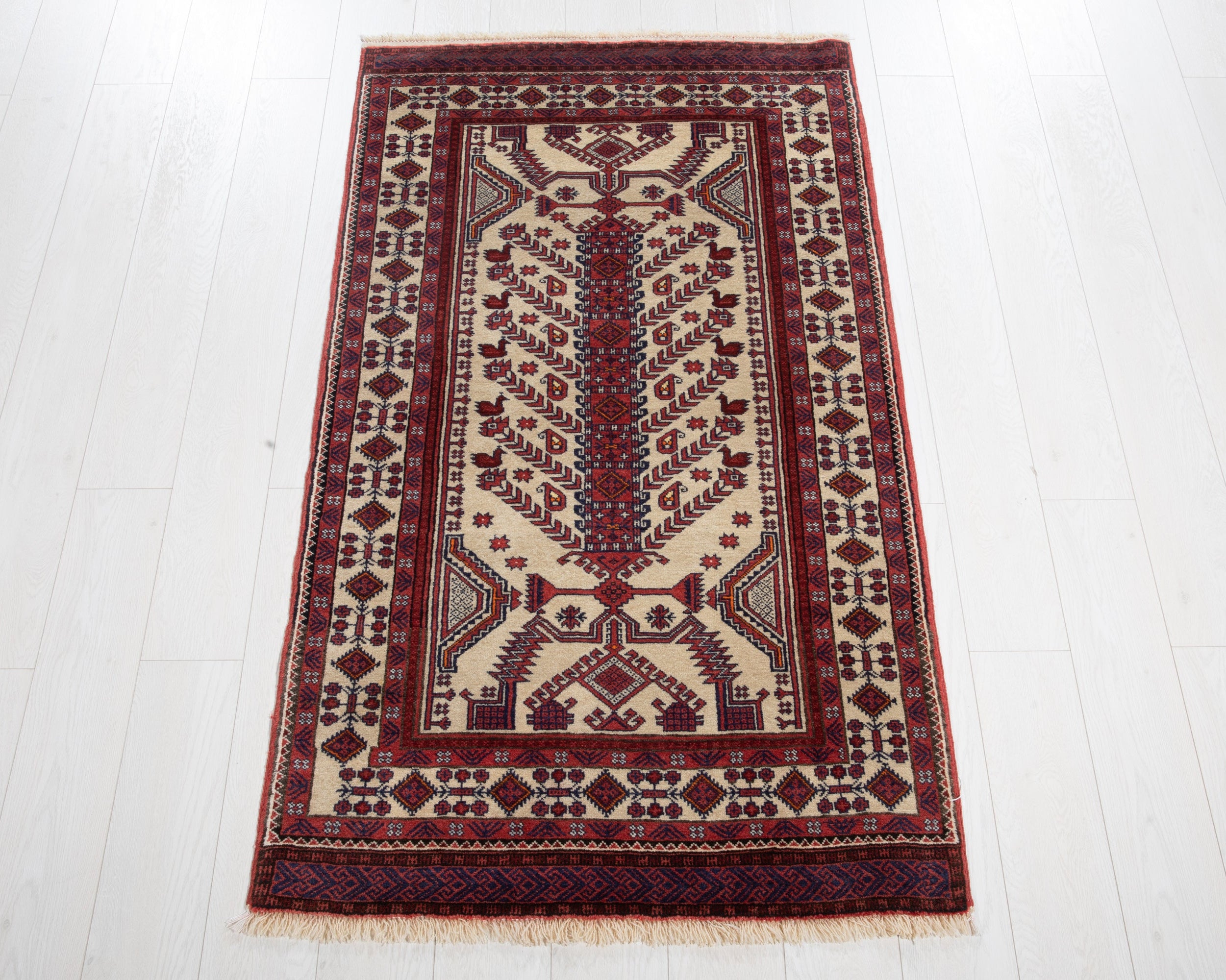 A traditional rectangular area rug with a central tree motif, featuring deep red, navy blue, and beige colors, laid out on a light-colored wooden floor.
