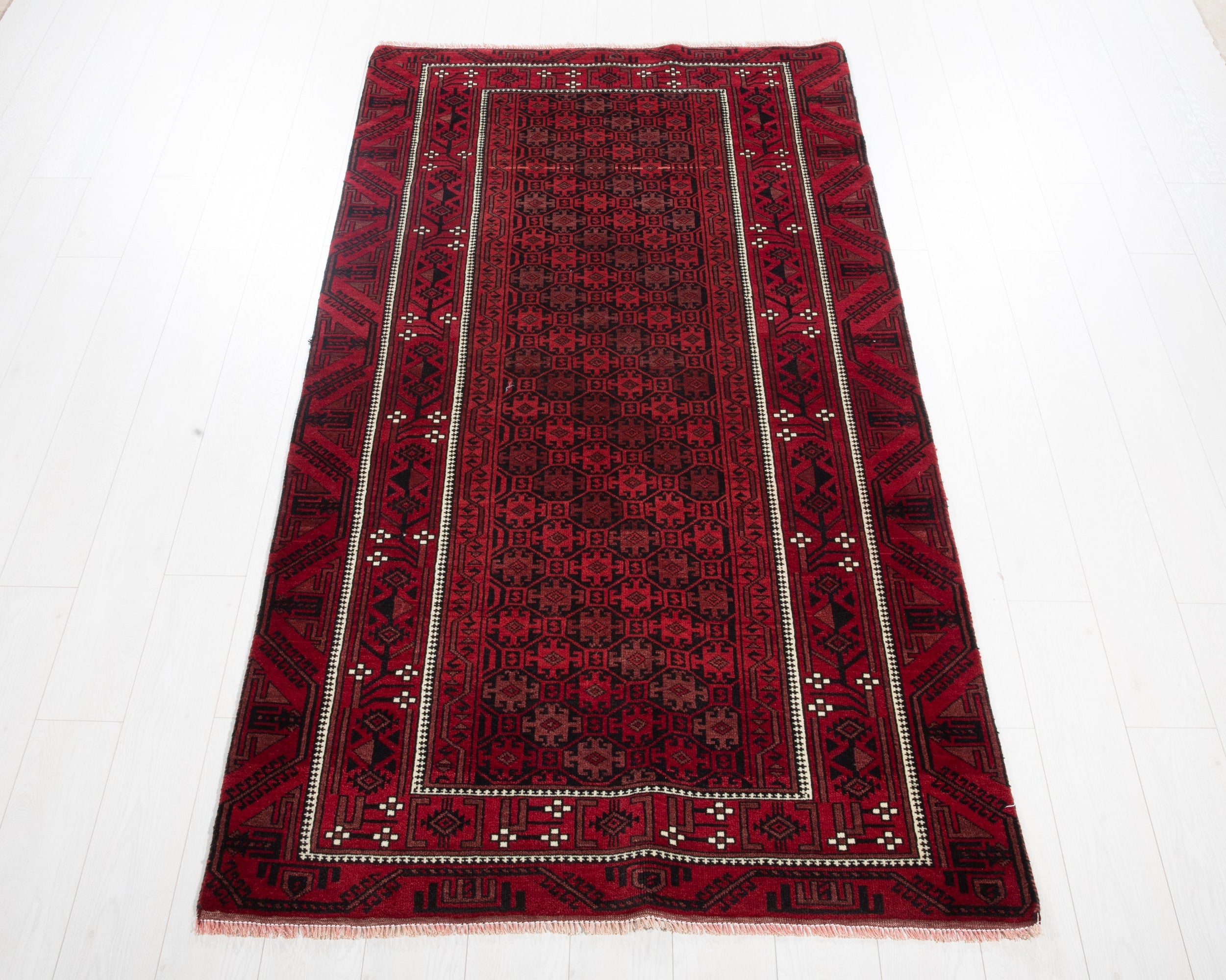 A rectangular red and black patterned rug laid flat on a white floor. The rug features a central field with intricate geometric motifs, bordered by red and black bands with cream-colored accents.