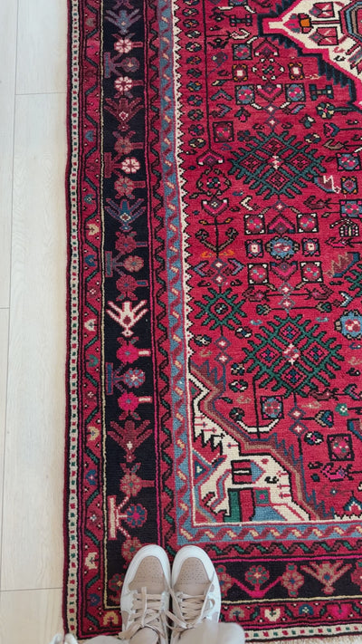 Close-up of Hand-Knotted Vintage Persian Malayer Red Soft Rug corner showing intricate floral border and geometric motifs beside a pair of shoes.