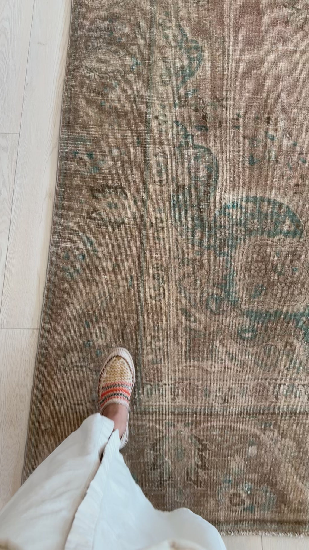 Close-up of the corner of a hand-knotted vintage wool rug with intricate faded teal and beige patterns, showing texture and foot for scale.